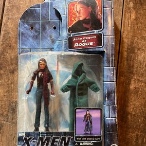 May include: An X-Men: The Movie action figure of Rogue, featuring Anna Paquin. The figure is in its original packaging, with a green cloak and scarf. The packaging includes the Marvel and Toy Biz logos.