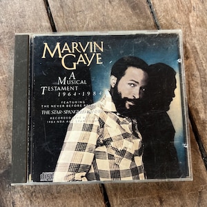 May include: A compact disc case featuring Marvin Gaye's album, "A Musical Testament 1964-1984." The cover art displays a portrait of Gaye in a plaid shirt against a blue background. The CD case is in a clear plastic case.