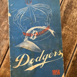 May include: A vintage 1956 Dodgers baseball official program and score card. The blue cover features a white line drawing of a crown topped with a baseball, the text World Champions, the team name Dodgers, and the year 1956 in orange lettering.