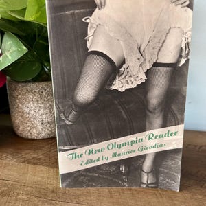 May include: Black and white book cover titled "The New Olympia Reader" edited by Maurice Girodias. The cover features a vintage photograph of a person wearing fishnet stockings and high heels.