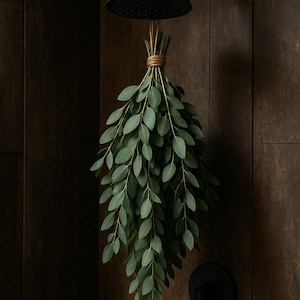 May include: A bundle of fresh eucalyptus leaves hangs from a black shower head. The leaves are a vibrant green, tied together with twine. The shower fixtures and the wall are a dark brown color.