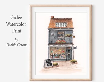 May include: A giclée watercolor print by Debbie Cerone featuring a dark gray two-story bookstore with a brown roof, a storefront window displaying colorful books, a hanging lantern, flower boxes, and a sidewalk sign that reads A book is a gift.