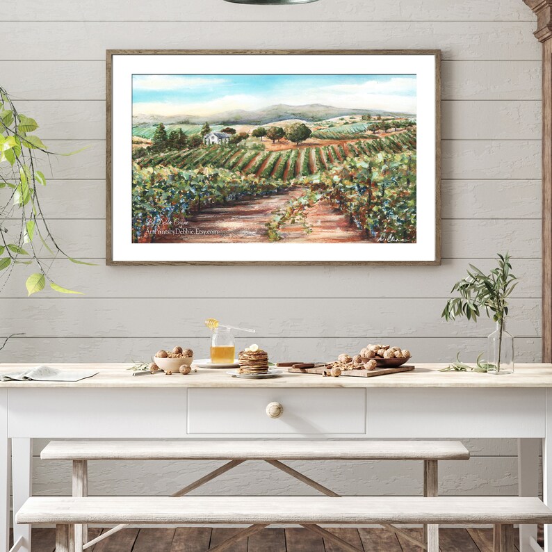 May include: A watercolor painting of a vineyard with a white farmhouse in the distance. The painting is framed in a light wood frame and hangs on a white wall.
