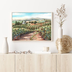 May include: A watercolor painting of a vineyard with rows of grapevines, a small white farmhouse, and rolling hills in the background. The painting is framed in a light wood frame.