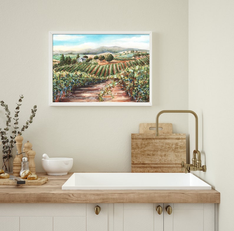 May include: A watercolor painting of a vineyard landscape with a white farmhouse in the distance. The painting is framed in a white frame and hangs on a light beige wall.