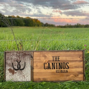 May include: A wooden sign with a rustic finish, featuring the text "THE CANINOS EST. 09.21.24" in black lettering. The sign is divided into two sections, with the left side showing a distressed white background with two black horseshoes. The sign is displayed against a backdrop of green grass and a barbed wire fence.