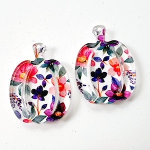 May include: Two clear acrylic pumpkin-shaped pendants featuring a vibrant floral pattern. The design includes various flowers in shades of pink, purple, orange, and dark blue, accented with green leaves, all set against a transparent background.