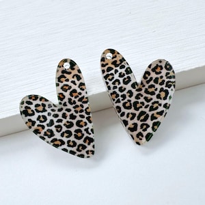 May include: Two heart-shaped acrylic charms with a leopard print pattern. The charms are tan with black spots and have a small hole at the top for hanging. They are displayed on a white surface.