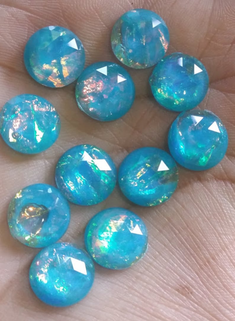 May include: A collection of round, faceted, light blue gemstones with iridescent flashes of color. The gemstones have a polished, reflective surface and are arranged in a scattered pattern. These gemstones would be suitable for jewelry making.
