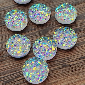 May include: A collection of iridescent, round cabochons with a druzy-like appearance. The cabochons are a mix of colors, including blue, yellow, and purple, and are set against a white base. They are arranged on a brown wooden surface.