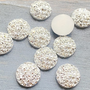 May include: A collection of round, silver-colored embellishments with a sparkling, textured surface. One white, smooth, round embellishment is also present. These decorative elements are likely used for crafts or jewelry making.