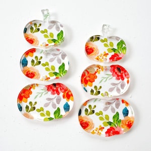 May include: Two sets of three stacked clear acrylic pumpkin-shaped pendants, each featuring a vibrant floral pattern with red, orange, and yellow flowers, green leaves, and grey foliage accents. The pendants have a smooth, glossy finish.