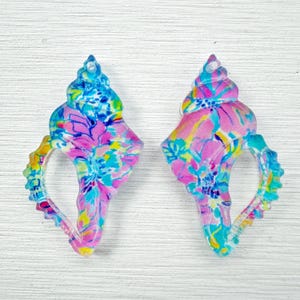 May include: Two seashell-shaped earrings with a colorful floral pattern in shades of pink, blue, yellow, and green. The earrings have a translucent appearance and a textured surface, with a small hole at the top for hanging.