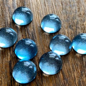 May include: Nine blue glass cabochons with a domed shape are arranged on a wooden surface. The cabochons have a gradient of blue hues, with a lighter, brighter blue at the base and a darker, more opaque blue at the top.
