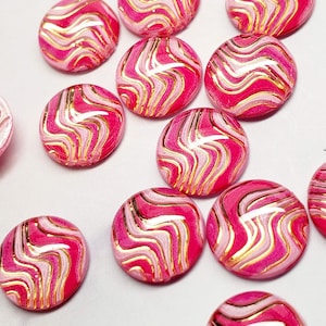 May include: A collection of pink, white, and gold swirl-patterned cabochons. The cabochons are round and have a glossy finish. The design features a vibrant pink base with white and gold metallic swirls.