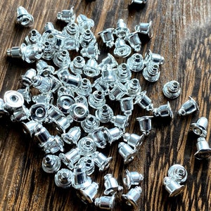 May include: A collection of small, shiny silver earring backs scattered on a dark wood surface. The earring backs have a bell-like shape with a flat, circular base and a post for securing earrings.