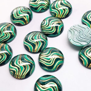 May include: A collection of round, decorative cabochons with a glossy finish. The cabochons feature a dark teal base with gold and light teal wavy lines. One cabochon has a light teal, textured surface. These are likely used for jewelry making or crafts.