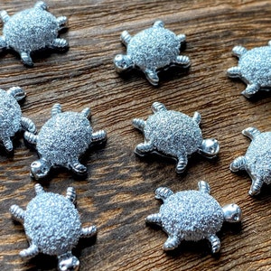 May include: A collection of silver-colored turtle charms with textured shells and detailed limbs. The charms are scattered on a wooden surface, showcasing their three-dimensional design. These decorative charms are ideal for jewelry making or craft projects.