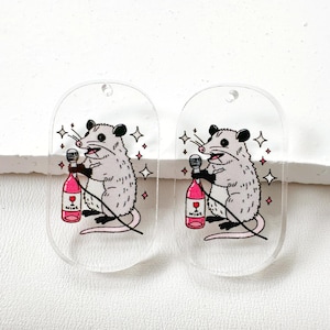 May include: Two oval-shaped clear acrylic pendants featuring a cartoon illustration of a gray opossum holding a pink wine bottle with the word WINE printed on the label. The opossum is also holding a microphone, surrounded by small star accents.