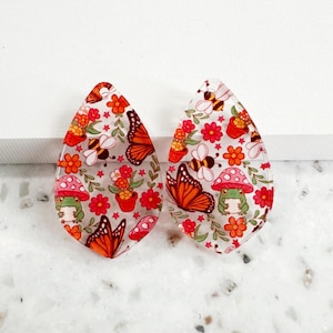 May include: A pair of teardrop-shaped acrylic pendants featuring a repeating pattern of orange monarch butterflies, green frogs with red mushroom caps, yellow and black bees, potted plants, and small red flowers on a clear background.