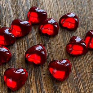 May include: A collection of thirteen red glass heart-shaped beads on a wooden surface.