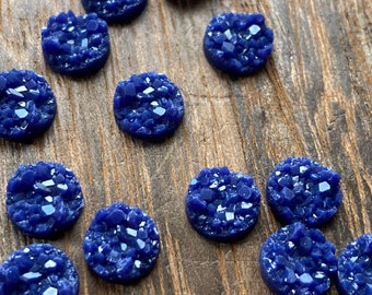 May include: A collection of 11 small, round, blue cabochons with a crystal-like texture. The cabochons are arranged in a semi-circle on a wooden surface.
