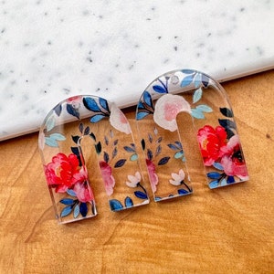 May include: Two clear acrylic arch-shaped earrings with a floral print. The print features pink, red, blue, and white flowers on a clear background.