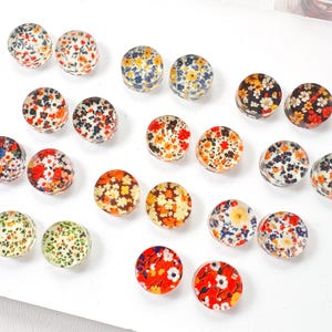 May include: A collection of circular cabochons featuring various floral patterns in vibrant colors including red, orange, yellow, blue, and green. Each piece has a smooth, glossy, domed surface with intricate, small-scale flower prints arranged in pairs.