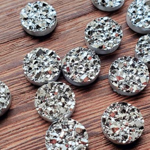 May include: A collection of silver-colored, round druzy cabochons with a sparkling, crystalline surface. The cabochons are set on a light gray base and arranged on a wooden surface. These are suitable for jewelry making and crafts.