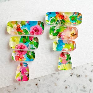 May include: A collection of rectangular and teardrop-shaped resin pieces, each showcasing a vibrant floral design. The flowers feature pink, red, blue, and yellow hues with green leaves, set against a clear background. These decorative pieces are ideal for jewelry making.