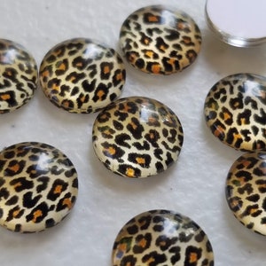 May include: A collection of round glass cabochons with a leopard print design. The cabochons feature a tan background with black and orange spots, and are suitable for jewelry making or crafting.