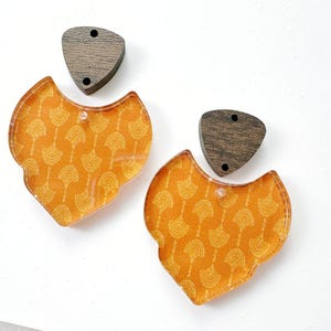May include: A pair of earrings featuring dark brown wooden triangular tops with two small holes and dangling orange acrylic shield-shaped pendants. The orange pendants display a repeating white fan-like pattern across their surfaces.