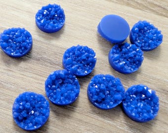 May include: A collection of round, cobalt blue cabochons with a druzy crystal effect. The cabochons are arranged on a light brown wooden surface. One cabochon is smooth, while the others have a textured, sparkling appearance.
