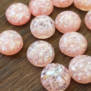 May include: A selection of round, pink-peach cabochons with a glittery, speckled appearance. The cabochons are scattered on a wooden surface. These decorative elements could be used for jewellery making or craft projects.