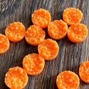 May include: A collection of orange, round, druzy-style cabochons. Each cabochon has a textured, crystalline surface and a flat back. The cabochons are arranged on a dark, wooden surface. These are suitable for jewelry making and crafting.