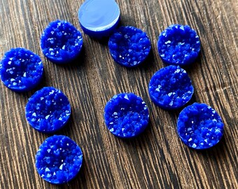May include: A collection of blue druzy cabochons with a sparkling, crystalline texture. The cabochons are round and vary slightly in size, with a smooth, polished surface. The background is a dark wood grain.