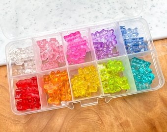 May include: A clear plastic storage box filled with colorful gummy bear shaped charms. The charms are arranged in rows by color: red, orange, yellow, green, blue, purple, pink, and clear.