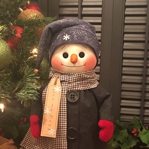May include: A snowman wearing a blue hat with a snowflake, a black coat with buttons, a black and white checkered scarf, and red mittens. The snowman is holding a tag that says "Let it Snow".