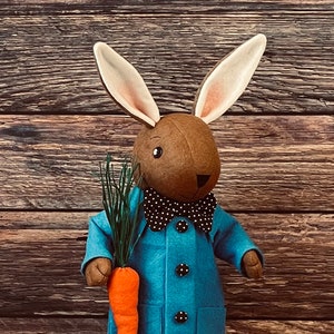 以下が含まれることがあります： 黒い水玉模様のボタンと黒白の水玉模様の蝶ネクタイをつけた青いコートを着た茶色のフェルトのウサギ。ウサギはニンジンと緑の草を持っています。