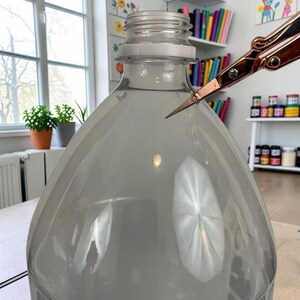 Pu&ograve; includere: Una bottiglia di plastica trasparente con un collo filettato viene tagliata da un paio di forbici in metallo color oro rosa. La bottiglia presenta una spalla liscia e arrotondata e un corpo ampio, con le forbici posizionate vicino al bordo.