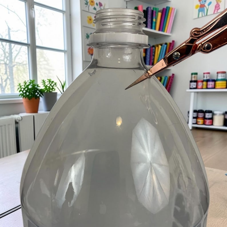 Pu&ograve; includere: Una bottiglia di plastica trasparente con un collo filettato viene tagliata da un paio di forbici in metallo color oro rosa. La bottiglia presenta una spalla liscia e arrotondata e un corpo ampio, con le forbici posizionate vicino al bordo.