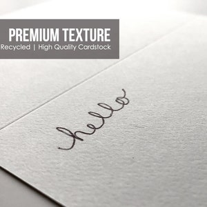 May include: A close-up of a white card with a textured surface. The word "hello" is written in black cursive handwriting.