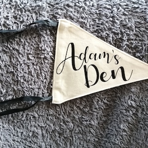 May include: Cream pennant flag with the words "Adam's Den" in black script. The triangular flag has black ribbons for hanging. The background is a textured grey surface.