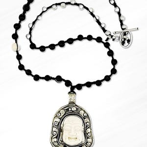 May include: A black beaded necklace with a silver toggle clasp and a silver pendant featuring a white carved Buddha face.
