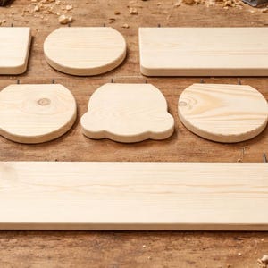 May include: A collection of unfinished wooden shapes, including squares, circles, and irregular forms, arranged on a wooden surface. The pieces vary in size and shape, with visible wood grain. Sawdust is scattered around the edges.