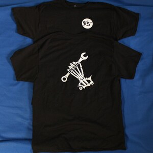 May include: Two black t-shirts, one stacked on top of the other. The top shirt has a small white circle with the letters "BS" inside. The bottom shirt has a white graphic of a skeleton hand holding a wrench.