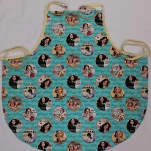 May include: A light blue apron with a repeating pattern of vintage Kellogg's Corn Flakes advertisements. The apron features a rounded bottom, yellow ties, and a yellow trim along the edges. The advertisements feature women and the words "Flavor" and "Drink".