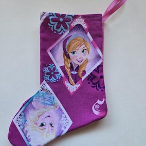 May include: A purple fabric Christmas stocking featuring a repeating diamond pattern with printed illustrations of two characters with blonde and braided hair. The stocking includes a pink ribbon loop at the top for hanging and blue snowflake accents.