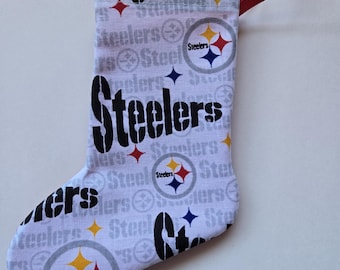 May include: A small, fabric Christmas stocking featuring the Pittsburgh Steelers logo and wordmark. The stocking is white with a repeating pattern of the team's logo and name. It has a red ribbon loop for hanging.