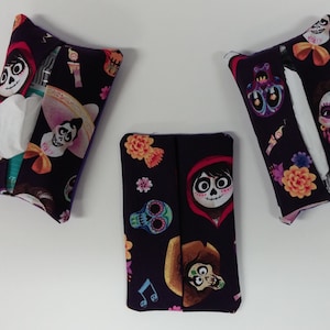 May include: Three purple fabric tissue box covers with a black background and colorful images of characters from the movie Coco. The characters include Miguel, Hector, and Dante. The covers are decorated with flowers, skulls, and musical notes.